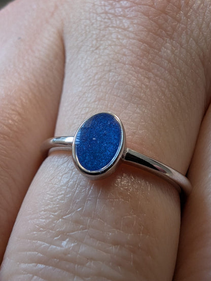 A sterling silver ring with an oval blue resin stone, worn on a person's finger. The stone includes ashes from a loved one.