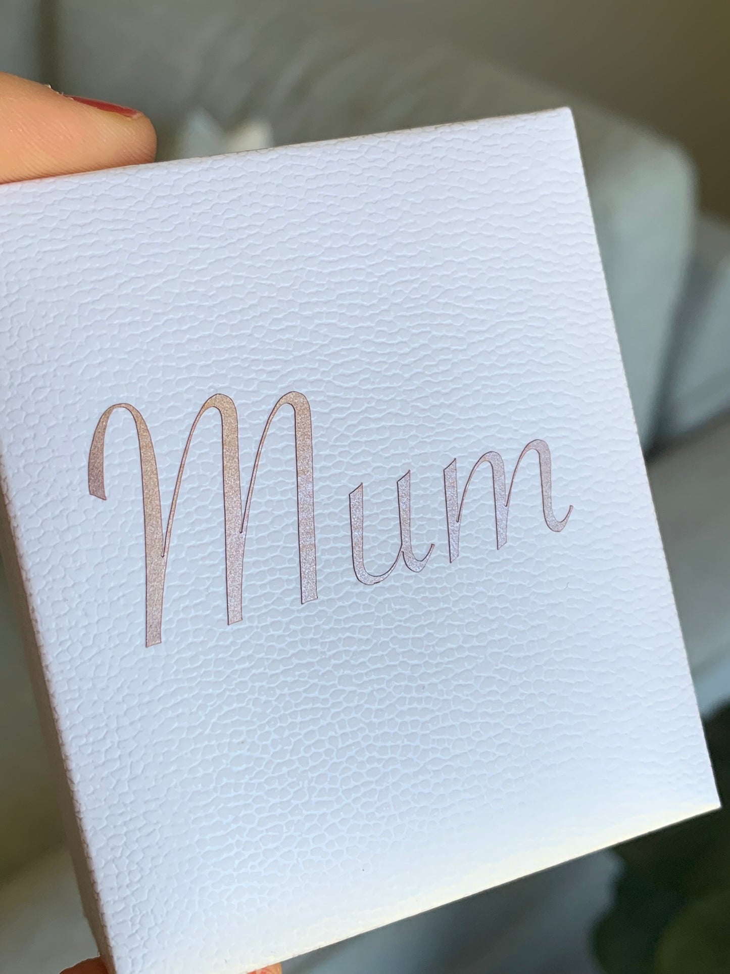 A close-up of a white textured gift box held by a hand, featuring the word 'Mum' printed in a metallic rose gold script font on the front.