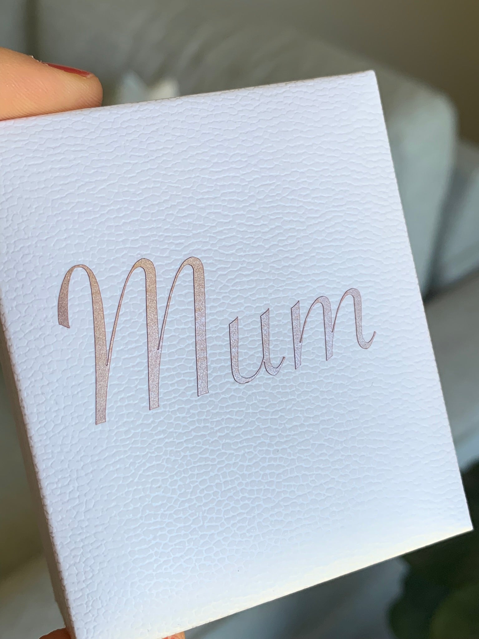 A close-up of a white textured gift box held by a hand, featuring the word 'Mum' printed in a metallic rose gold script font on the front.