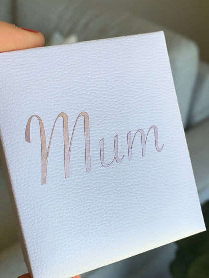 A close-up of a white textured gift box held by a hand, featuring the word 'Mum' printed in a metallic rose gold script font on the front.
