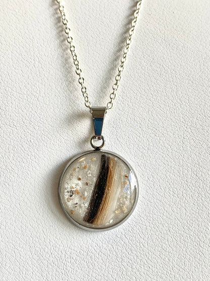 A silver chain necklace with a round pendant featuring a natural brown and black ashes and hair inside a clear resin dome, displayed on a textured white background.